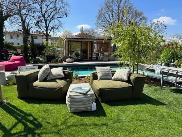 Outdoor seating area with two green beanbag chairs, a table with magazines, and a pool. Trees, a shed, and a house are in the background.