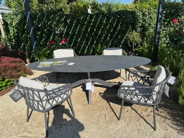 A gray outdoor table with four chairs is set up in a garden with a hedge, plants, and flowers.