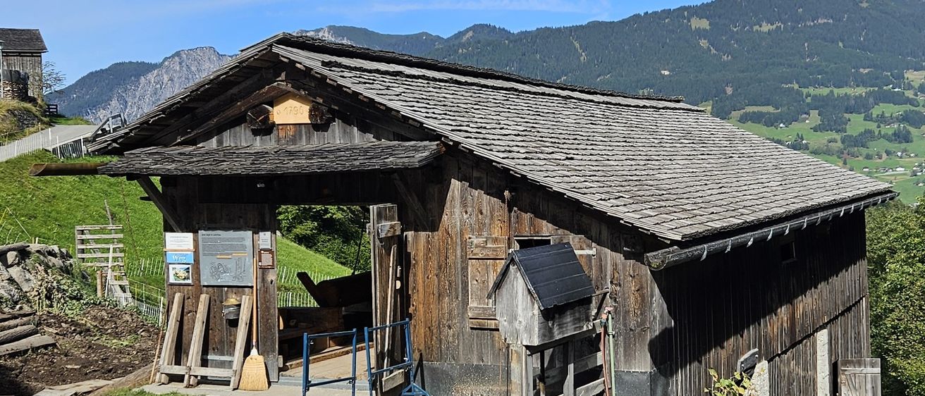 Ein altes Holzgebäude mit einem Schieferdach, in einem Gebirgsgebiet gelegen. Ein Schild über dem Eingang trägt die Aufschrift '1790'. Draußen stehen ein Besen und ein Eimer.
