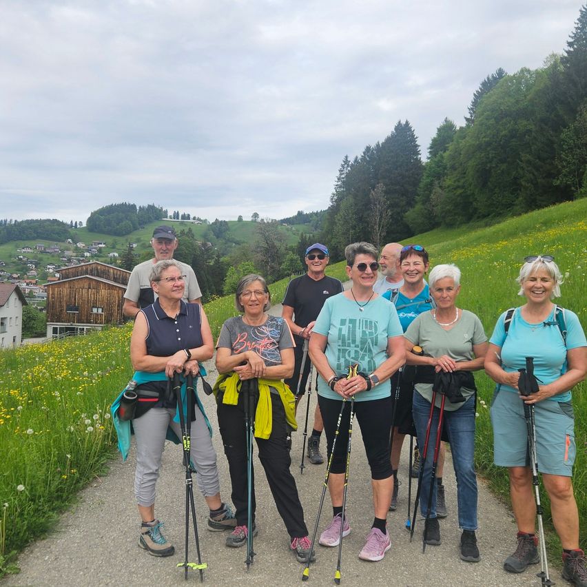 Eine Gruppe von Wanderern posiert für ein Foto auf einem Bergpfad, mit Häusern und Bäumen im Hintergrund. Sie alle lächeln und halten Wanderstöcke.
