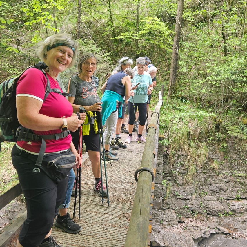 Eine Gruppe lächelnder Wanderer, ausgestattet mit Rucksäcken und Trekkingstöcken, geht über eine Holzbrücke in einem üppigen Wald.