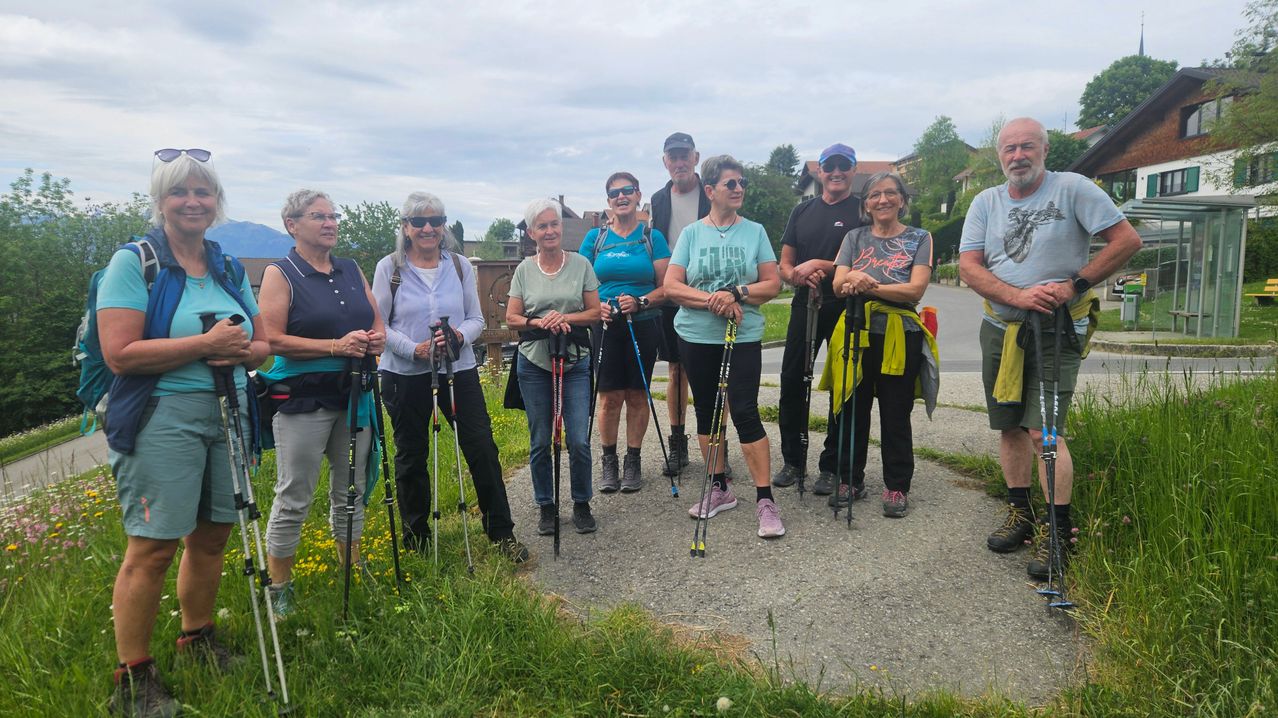 Eine Gruppe von Wanderern steht auf einem Kiesweg, lächelt und hält Trekkingstöcke. Im Hintergrund ist ein wolkiger Himmel und einige Häuser zu sehen.
