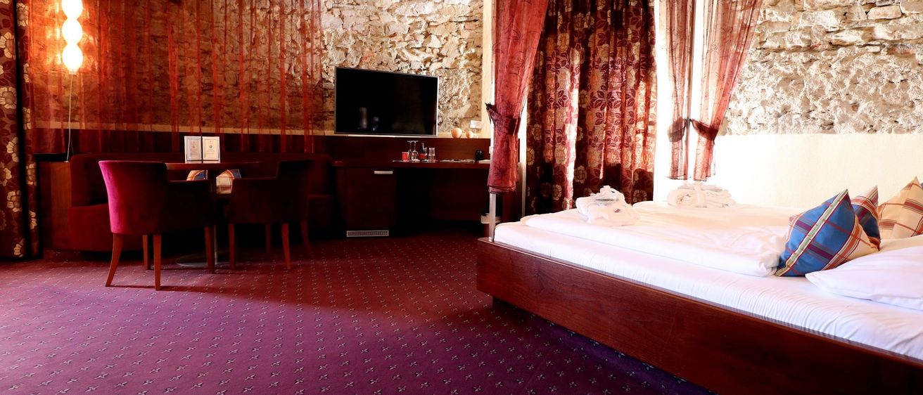 Ein Hotelzimmer mit Bett, Schreibtisch und Fernseher an einer Steinwand. Das Bett hat weiße Laken und einen braunroten Vorhang.