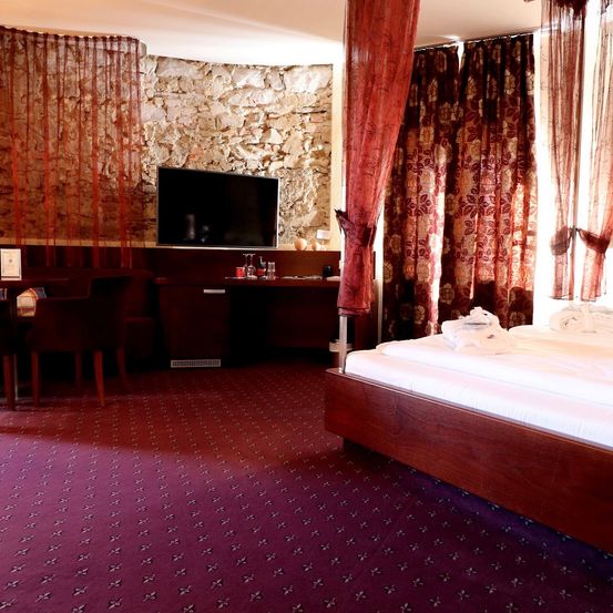 Ein Hotelzimmer mit Bett, Schreibtisch und Fernseher an einer Steinwand. Das Bett hat eine weiße Bettdecke und einen braunen Vorhang.