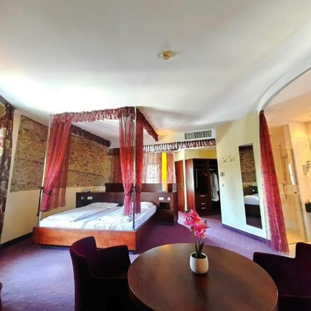 Ein gemütliches Hotelzimmer mit einem Himmelbett, einem runden Tisch mit Blumen und zwei Stühlen. Das Zimmer hat eine Steinwand, einen Kleiderschrank und einen Duschbereich.