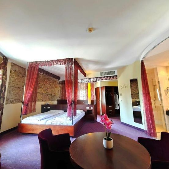 Ein gemütliches Hotelzimmer mit einem Himmelbett, einem runden Tisch mit Blumen und zwei Stühlen. Das Zimmer hat eine Steinwand, einen Kleiderschrank und einen Duschbereich.