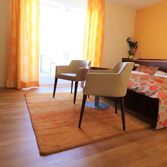 Ein Schlafzimmer mit Holzböden, einem Bett mit gemusterter Decke, zwei Stühlen, einem kleinen Tisch und einer Pflanze auf dem Kopfteil. Das Zimmer hat ein Fenster mit gelben Vorhängen.