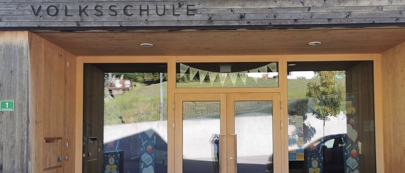 Der Eingang eines hölzernen Gebäudes namens 'Volksschule' mit zwei Glastüren, die einen Hügel, einen Baum und ein Auto draußen reflektieren.