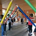 Eine Gruppe von Kindern und Erwachsenen hält farbige Buntstifte in einem Raum und bildet einen Kreis. Sie tragen Freizeitkleidung. Eine Uhr ist an der Wand angebracht.
