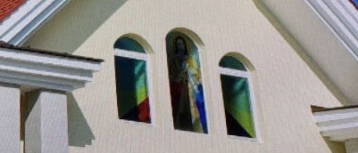 Die Fassade einer Kirche mit weißen Wänden und einem braunen Dach, geschmückt mit einem schwarzen Turm, der gewölbte Fenster und farbenfrohe Buntglasfenster mit religiösen Darstellungen aufweist.
