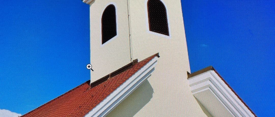 Eine weiße Kirche mit rotem Dach und schwarzem Turm. Drei bunte Fenster sind zu sehen, eines davon zeigt eine religiöse Figur.