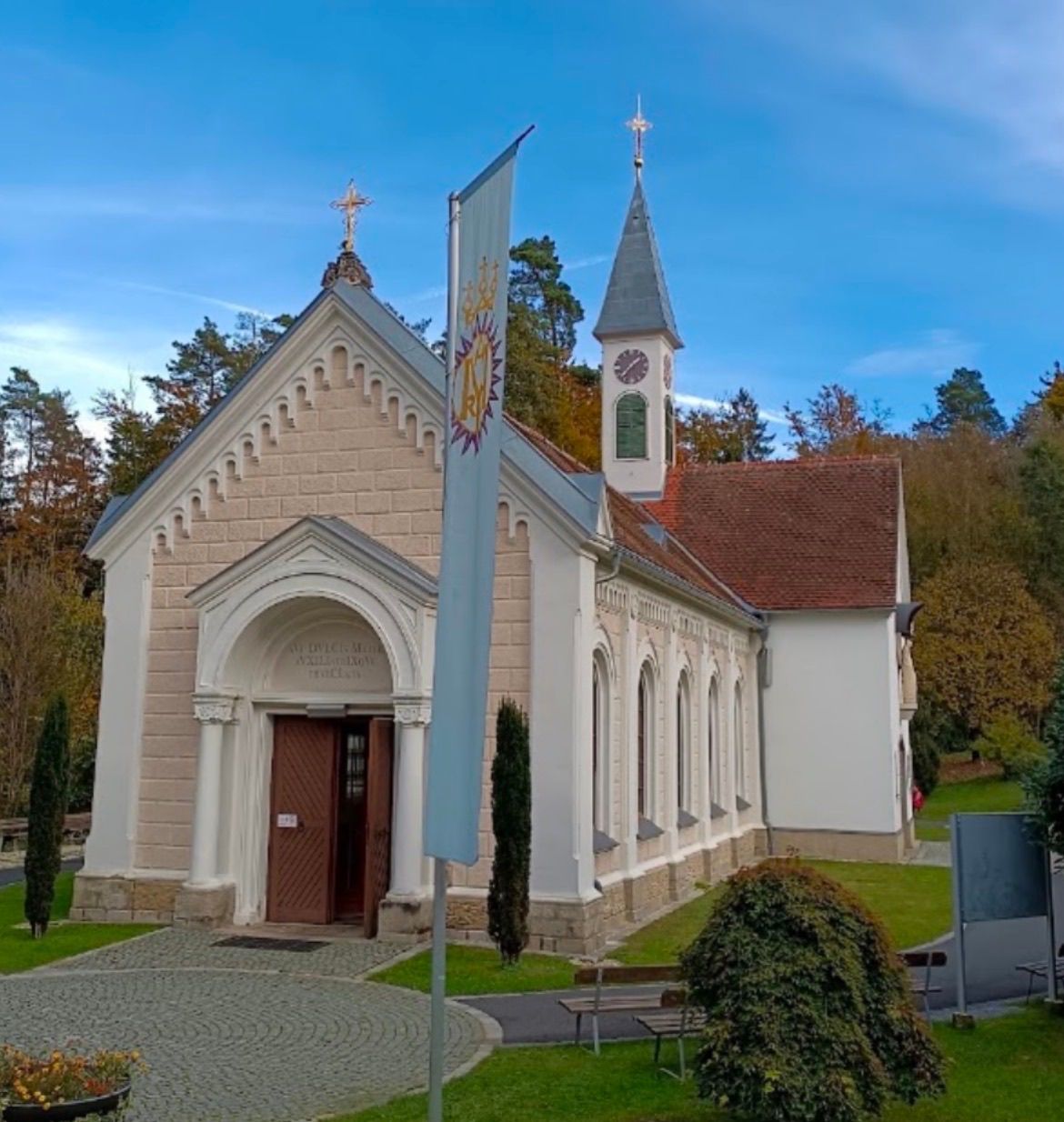 A small church with a brown roof and white walls, featuring a clock tower, a cross on top, and an arched entrance with a wooden door.