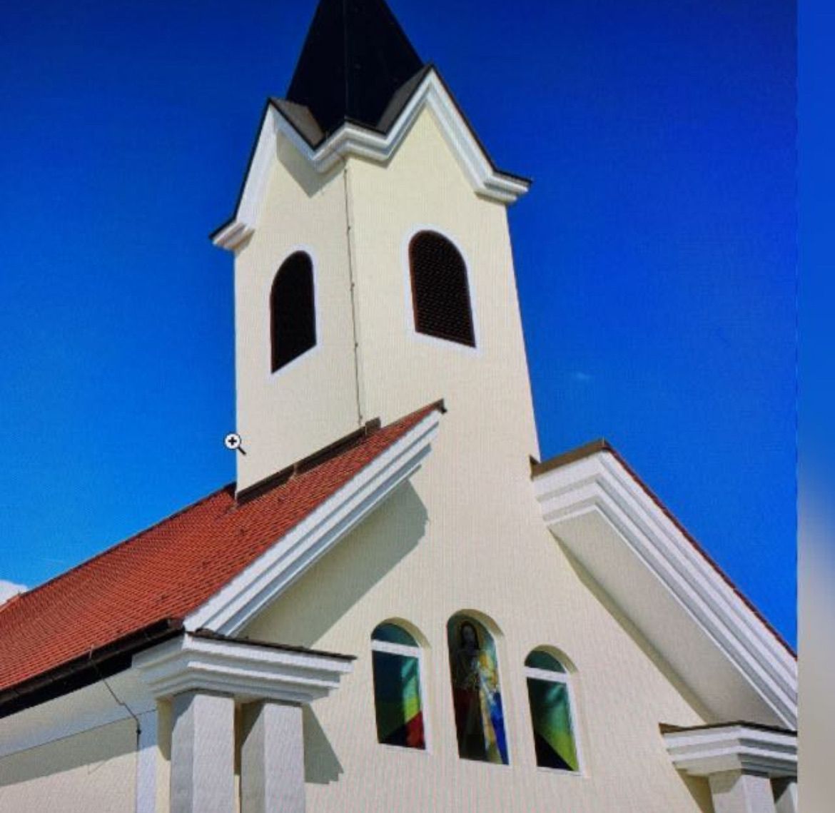 Eine weiße Kirche mit einem roten Dach und einem Kirchturm mit schwarzem Dach. Die Kirche hat drei Bogenfenster mit Buntglas.