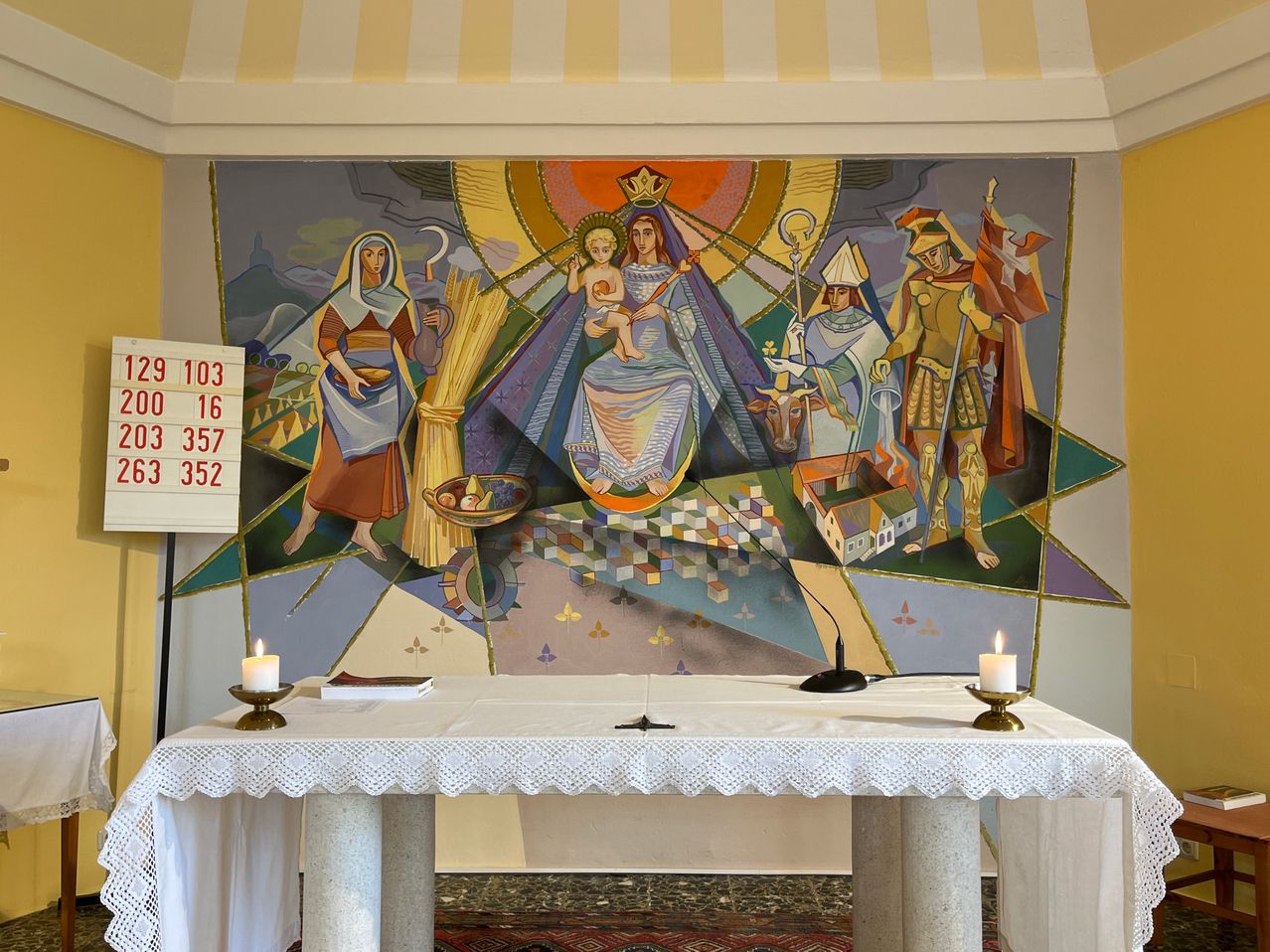 Ein Wandbild, das eine religiöse Szene mit Figuren wie einer Frau, einem Kind und anderen darstellt. Zwei Kerzen auf einem Tisch davor.