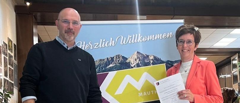 Ein Mann und eine Frau stehen vor einem Werbeplakat für die Marktgemeinde Kotschach-Mauthen. Die Frau hält ein Zertifikat.