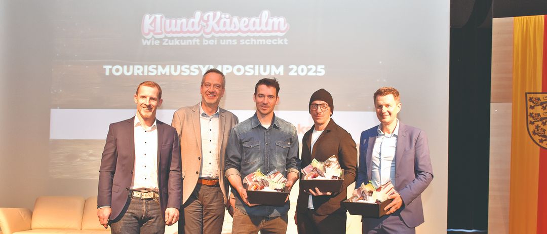 Fünf Männer stehen lächelnd auf einer Bühne und halten Boxen. Dahinter zeigt eine Leinwand 'Klund Kaisealm Wie Zukunft bei uns schmeckt' und 'TOURISMUSSYMPOSIUM 2025'. Eine Couch und eine Pflanze sind im Hintergrund.