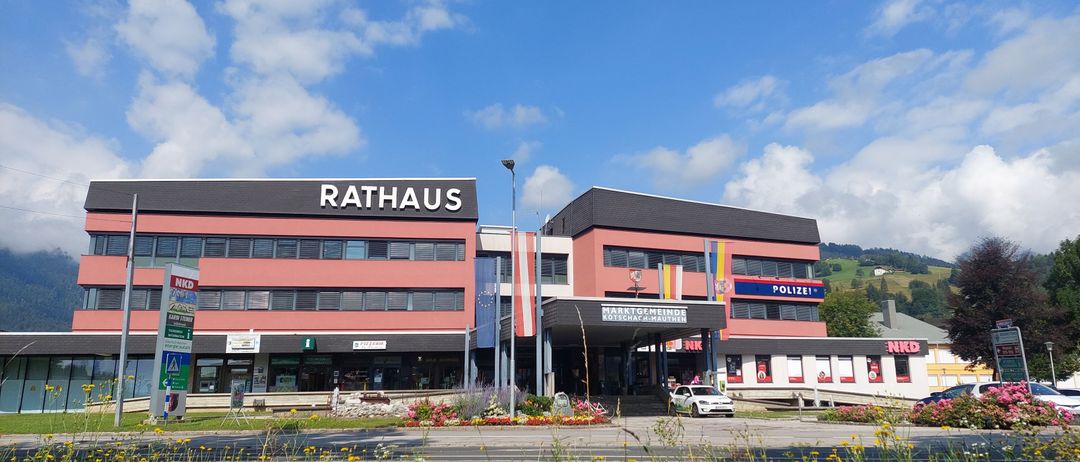 Ein Gebäude namens Rathaus mit einem Polizeischild davor. Ein weißes Auto ist davor geparkt. Vor dem Gebäude befinden sich Blumen und Gras.