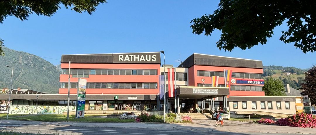 Das Rathausgebäude in Kotschach-Mauthen, Österreich, hat eine rote Fassade mit Fahnen. Es befindet sich in der Nähe einer Straße, vor der zwei Personen gehen.
