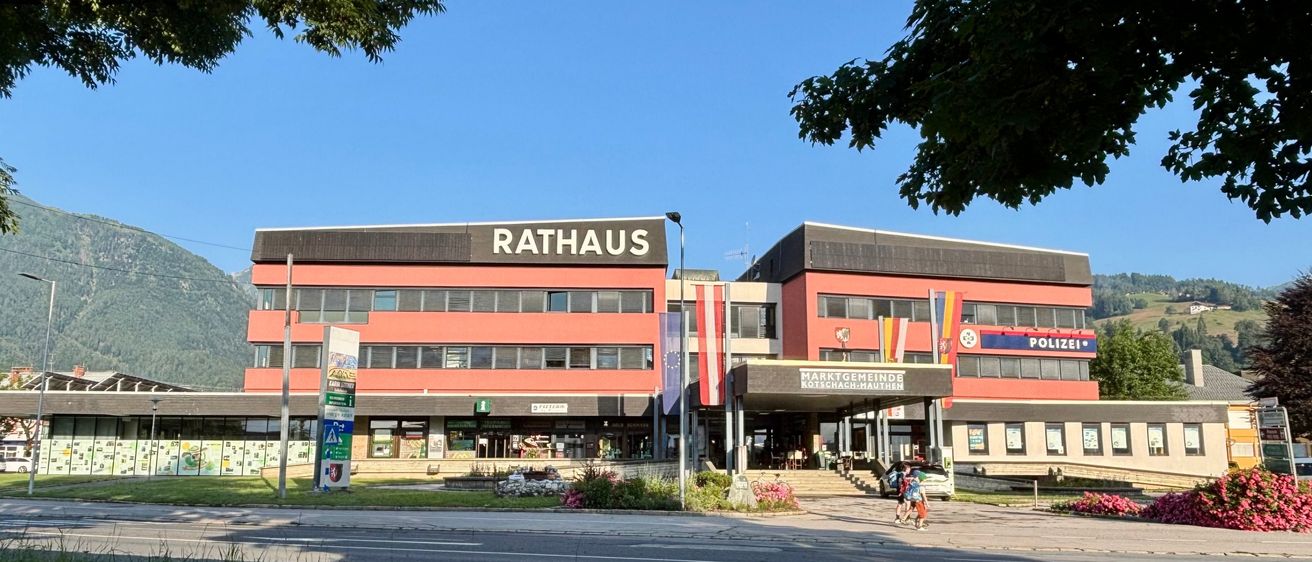 Das Rathausgebäude in Kotschach-Mauthen, Österreich, hat eine rote Fassade mit Fahnen. Es befindet sich in der Nähe einer Straße, vor der zwei Personen gehen.