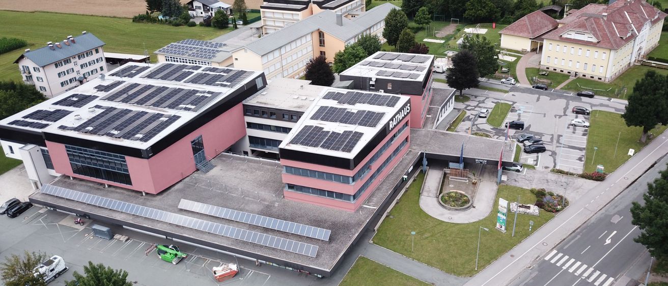 Ein Luftbild eines modernen Gebäudes mit Solarmodulen auf dem Dach, umgeben von einem Parkplatz, Bäumen und Gebäuden. Das Gebäude ist rosa und hat eine Flagge an seiner Seite.