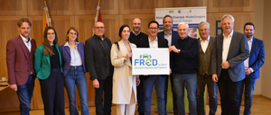 Eine Gruppe von Menschen steht gemeinsam und hält ein Schild mit der Aufschrift 'FROD'. Sie tragen formelle Kleidung. Dahinter ist eine Holzwand mit einer Flagge und einem Banner zu sehen.