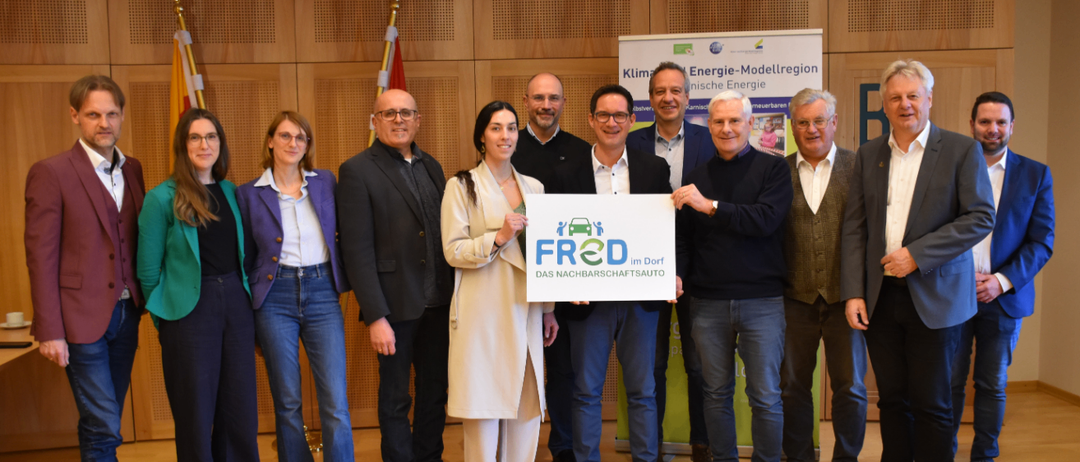 Eine Gruppe von Menschen steht gemeinsam und hält ein Schild mit der Aufschrift 'FROD'. Sie tragen formelle Kleidung. Dahinter ist eine Holzwand mit einer Flagge und einem Banner zu sehen.