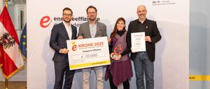 Vier Personen stehen vor einem Banner mit dem Text 'Land Kärnten' und einem Logo. Sie halten ein Zertifikat und einen Scheck mit dem Text 'Krone 2025' und dem Betrag von 10.000 Euro. Die Frau hält eine Trophäe. Sie tragen alle formelle Kleidung.
