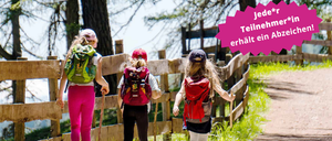 Drei Mädchen wandern mit ihren Rucksäcken, tragen Mützen und halten Trekkingstöcke. Sie gehen auf einem Erdweg in der Nähe eines Holzzauns.