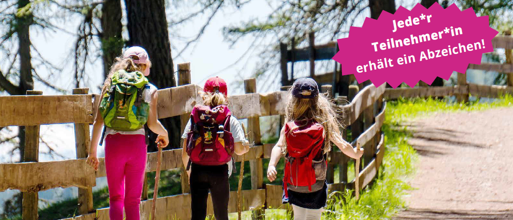 Drei Mädchen wandern mit ihren Rucksäcken, tragen Mützen und halten Trekkingstöcke. Sie gehen auf einem Erdweg in der Nähe eines Holzzauns.