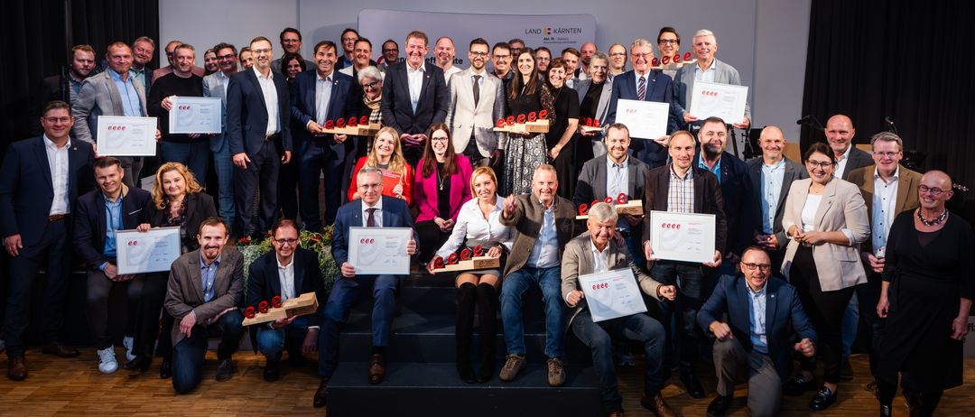 Eine Gruppe von Menschen in formeller Kleidung posiert für ein Foto. Sie halten Urkunden und Holzpreise in den Händen. Einige von ihnen sitzen auf dem Boden, während andere stehen. Sie befinden sich wahrscheinlich in einem Konferenzraum. Ein weißer Hintergrund ist hinter ihnen.