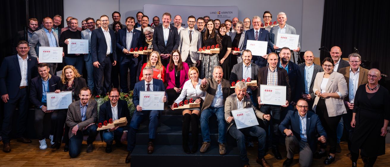 Eine Gruppe von Menschen in formeller Kleidung posiert für ein Foto. Sie halten Urkunden und Holzpreise in den Händen. Einige von ihnen sitzen auf dem Boden, während andere stehen. Sie befinden sich wahrscheinlich in einem Konferenzraum. Ein weißer Hintergrund ist hinter ihnen.