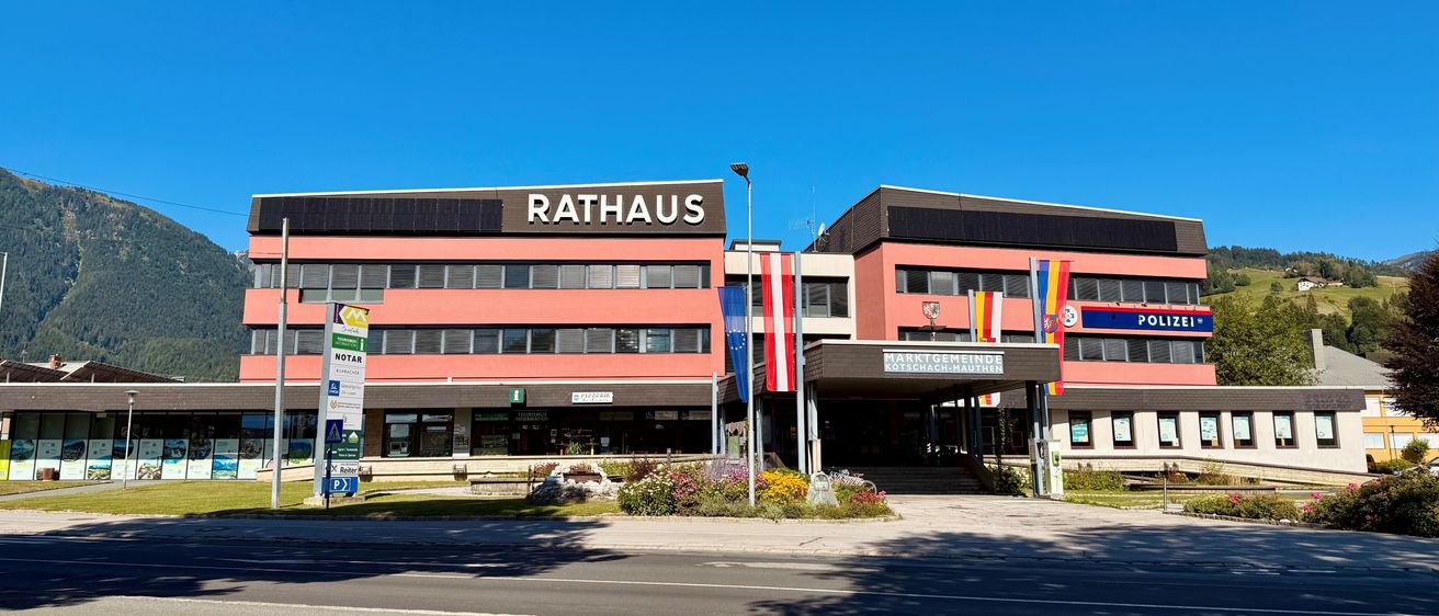 Das Rathaus-Gebäude ist prominent mit einem klaren blauen Himmel im Hintergrund zu sehen. Das Gebäude hat ein modernes Design mit Glasfenstern und einem großen Schild, das Rathaus steht. Flaggen sind an Stangen vor dem Gebäude angebracht.