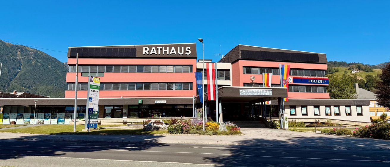 Das Rathausgebäude in Kotschach-Mauthen, Österreich, zeichnet sich durch ein modernes architektonisches Design aus, mit einem großen Schild, das Rathaus steht. Das Gebäude ist von Grünflächen umgeben und hat mehrere Flaggen, einschließlich der österreichischen Flagge, vor einem klaren blauen Himmel.