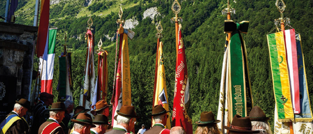 Eine Gruppe von Menschen in traditioneller Kleidung steht vor Flaggen mit einem Gebirgshintergrund.