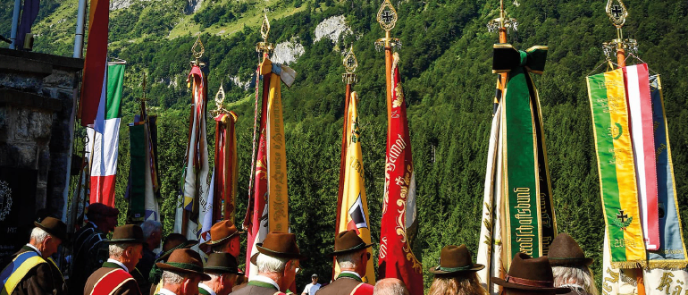 Eine Gruppe von Menschen in traditioneller Kleidung steht vor Flaggen mit einem Gebirgshintergrund.