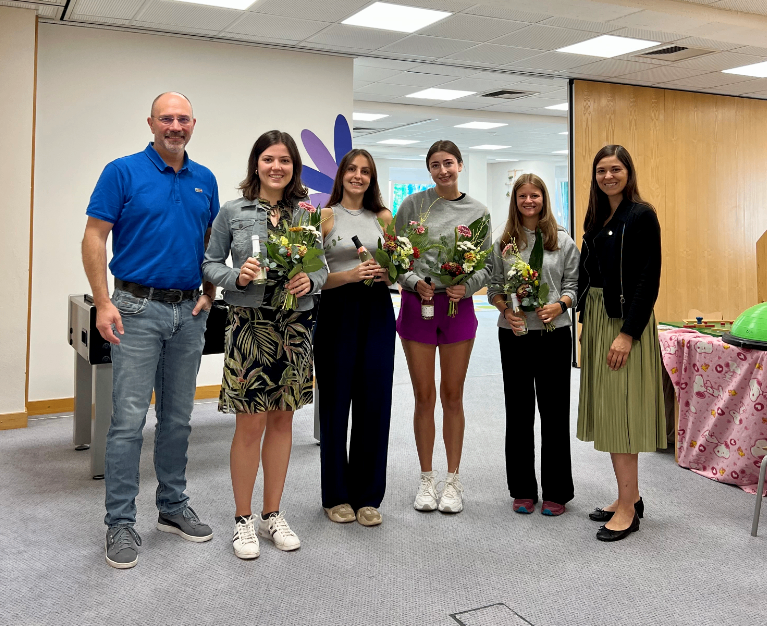 Eine Gruppe lächelnder Frauen mit Blumen posiert für ein Foto mit einem Mann in einem blauen Hemd in einem Raum mit weißen Wänden und grauem Teppich.