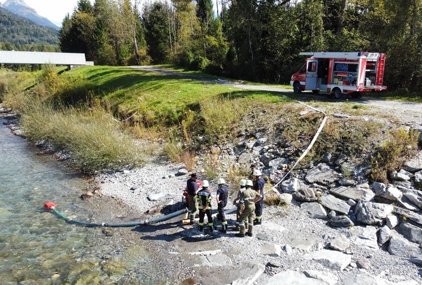 Feuerwehrleute verwenden eine Schlange in der Nähe eines Flusses, mit einem Feuerwehrwagen in der Nähe. Sie befinden sich auf felsigem Gelände mit einer Brücke im Hintergrund.