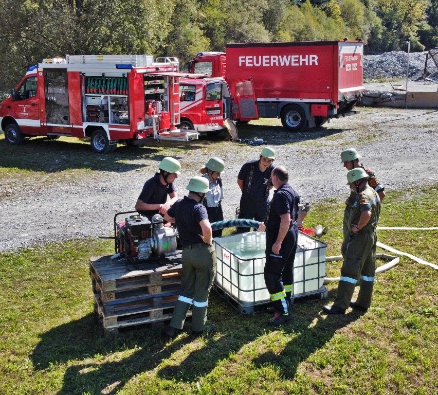 Eine Gruppe von Feuerwehrleuten in Uniform steht um einen Feuerwehrwagen herum. In der Nähe gibt es einen Wassertank, einen Schlauch und einen Generator. Sie befinden sich auf einer kiesigen Oberfläche mit Gras.