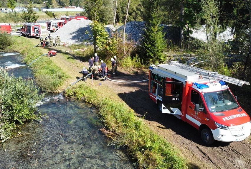 Ein Feuerwehrfahrzeug steht in der Nähe eines Flusses, wobei Feuerwehrleute eine Situation am Flussufer behandeln. Eine Leitung ist auf dem Boden verlegt.