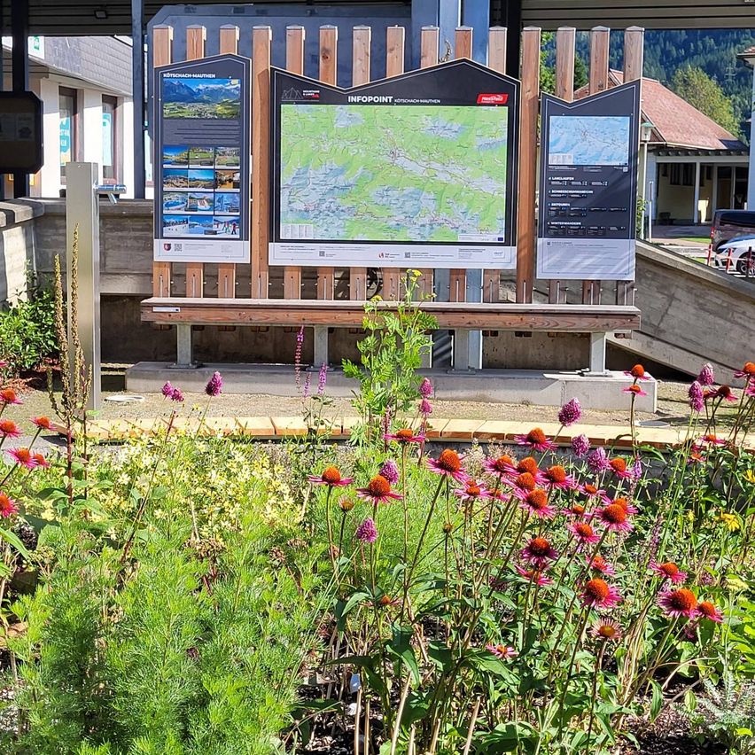 Ein Garten mit verschiedenen bunten Blumen und Pflanzen. Ein Holzzaun mit einem Schild, das 'INFOPOINT' und eine Karte zeigt. Im Hintergrund befindet sich ein Haus mit einem Dach.