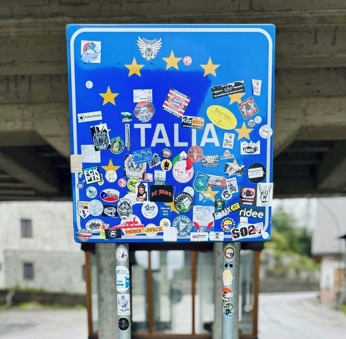 A blue sign covered in various stickers and labels stands under a bridge. The sign says 'ITALIA' prominently. There are stars, logos, and text stickers from different countries and brands.