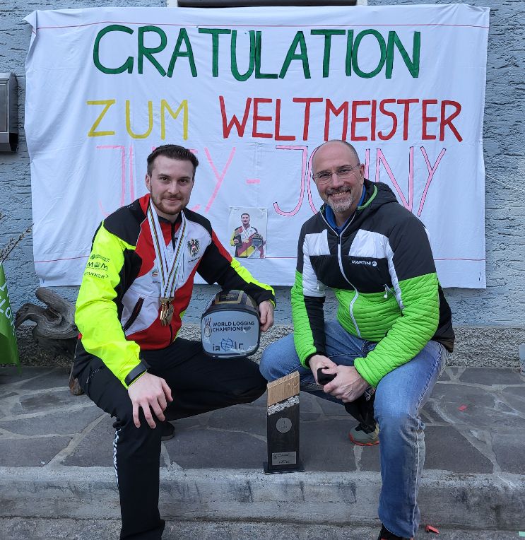 Two men sit on steps, one holds a helmet trophy, the other wears glasses and a jacket. Behind them is a banner that reads 'Congratulation zum Weltmeister'.
