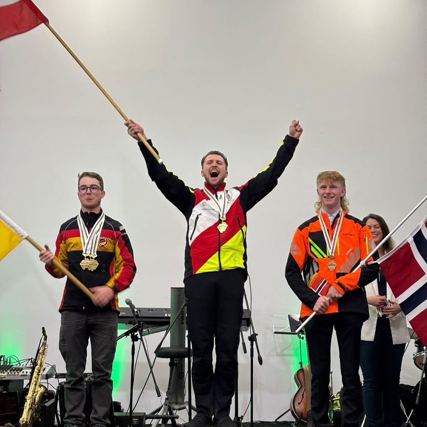 Three men wearing jackets and medals stand on a stage with flags. A man in the middle celebrates with raised arms, while others hold flags. Musical instruments are visible in the background.