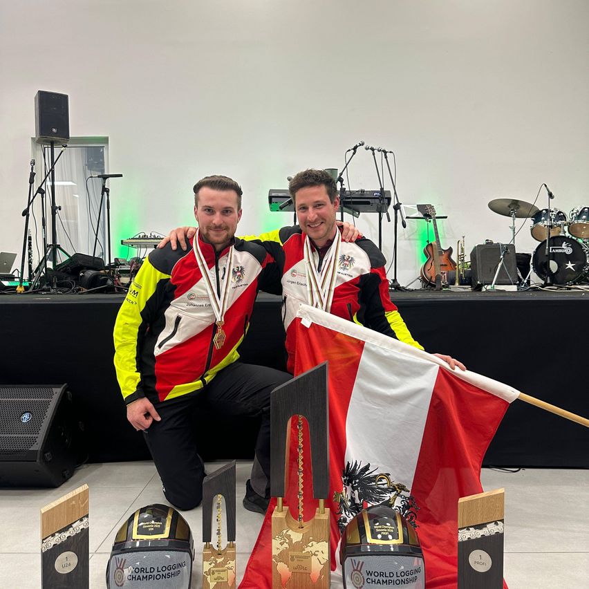 Two men kneel with trophies, medals, and a flag, smiling in front of a stage with musical instruments.