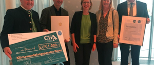 Fünf Personen in formeller Kleidung stehen zusammen und halten Urkunden und ein großes grünes Plakat mit einem Geldpreis. Der Hintergrund zeigt ein deutsches Regierungsbanner.