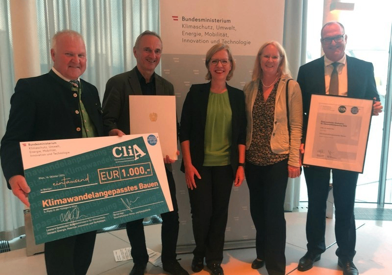 Fünf Personen in formeller Kleidung stehen zusammen und halten Urkunden und ein großes grünes Plakat mit einem Geldpreis. Der Hintergrund zeigt ein deutsches Regierungsbanner.