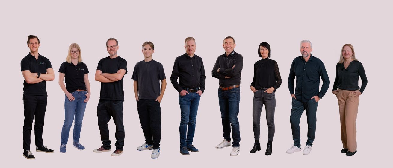 Fünf Personen, alle in Schwarz gekleidet, stehen in einer Reihe. Die beiden Männer auf der linken Seite haben ihre Arme verschränkt, während der Mann in der Mitte und die beiden auf der rechten Seite ihre Hände in den Taschen haben.