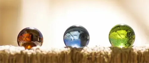 Drei farbige Glasmurmeln auf einer strukturierten Oberfläche. Die linke Murmel ist bernsteinfarben, die mittlere blau und die rechte grün.
