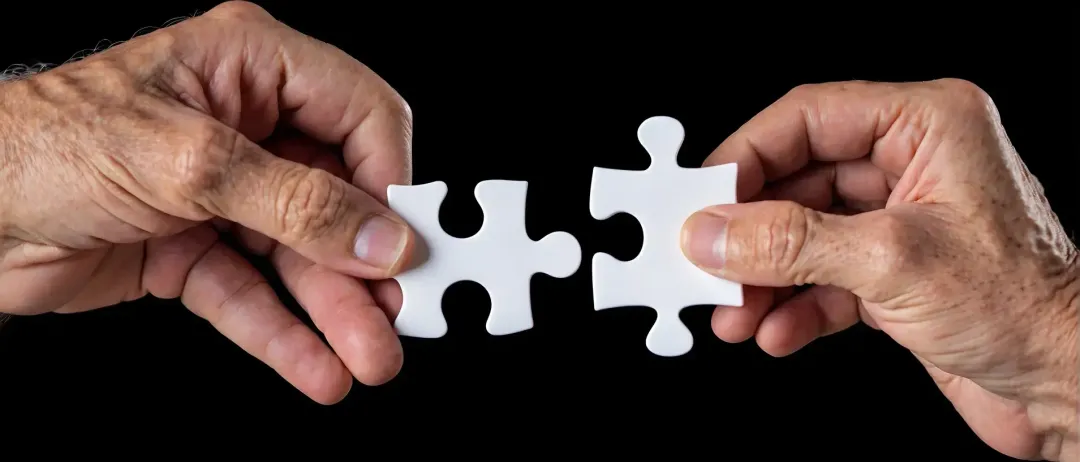 Zwei Hände halten ein weißes Puzzleteil vor schwarzem Hintergrund. Die Puzzleteile liegen nah beieinander und deuten auf eine Verbindung hin.