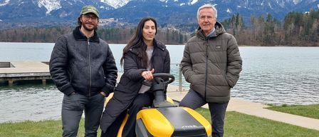 Drei Personen posieren in der Nähe eines Sees mit Bergen im Hintergrund. Der Mann links trägt eine Brille und einen Hut, die Frau in der Mitte hält eine Rasenmäher, und der Mann rechts lächelt.